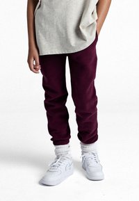 Un pantalon de survêtement bordeaux avec des poignets élastiques, associé à des baskets blanches. Le t-shirt est gris clair, de coupe décontractée avec un ourlet arrondi.