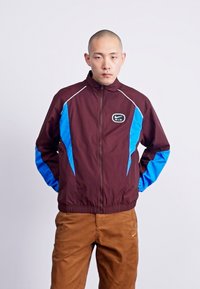 Vinröd vindjacka med blå accenter och vit piping, med dragkedja fram och en liten logotyp på bröstet. Strukturerad tyg.