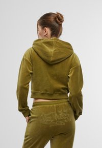 Haut à capuche en velours olive vert, coupé court, avec capuche et poignets côtelés. Présente un logo brodé sur le jogger assorti, une texture douce et une coupe décontractée.