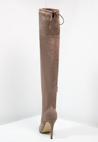 Bota de ante beige claro hasta el muslo con punta afilada y tacón de aguja delgado. Presenta cordones decorativos en la parte superior y detalles de costuras.