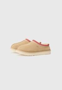 Beige suède instappers met een gestructureerde, rood-witte geweven accent langs de kraag en een crèmewitte rubberen zool.