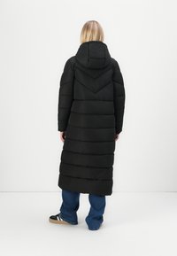 Lang zwarte donzen jas met een capuchon, voorzien van een chevronontwerp op de rug. Horizontale stiksels en een rechte silhouet.