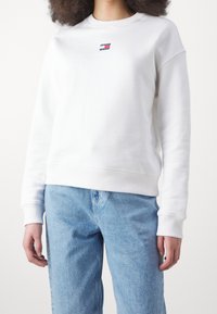 Persona che indossa una felpa bianca a maniche lunghe di Tommy Jeans e jeans di denim blu chiaro, in piedi di fronte a uno sfondo bianco semplice.