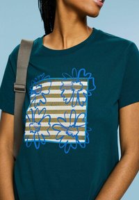 Teal bomullströja med ett stort blommönster i blått på en randig rektangulär bakgrund i guld- och vitnyanser. Kortärmad.