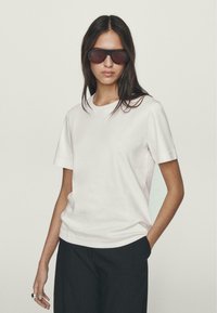 T-shirt bianca in cotone con maniche corte e scollatura rotonda, abbinata a pantaloni scuri e larghi. Il modello indossa occhiali da sole scuri di grandi dimensioni.