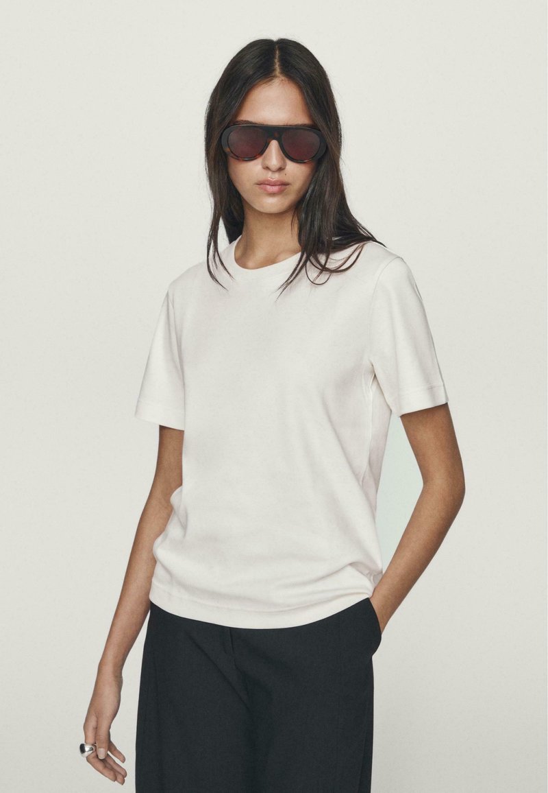 T-shirt bianca in cotone con maniche corte e scollatura rotonda, abbinata a pantaloni scuri e larghi. Il modello indossa occhiali da sole scuri di grandi dimensioni.