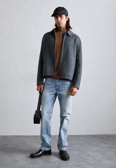 Graue Wolljacke, brauner Rollkragenpullover, hellblaue Jeans, schwarze Lederstiefel und eine schwarze Mütze; hält eine schwarze Umhängetasche.
