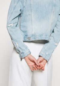 Veste en denim bleu clair avec une coupe courte, des accents usés et des détails de fermeture éclair en argent sur les manches. Portée avec un pantalon blanc, les mains croisées derrière.