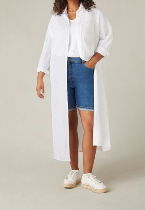 Žena v dlouhé bílé košili na knoflíky přes bílý top, modré džínové šortky, bílé tenisky a náhrdelník, stojící s rukou v kapse.