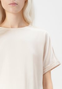 Beige kortermet topp med løs passform, rund halslinje og glatt tekstur. Ermer med en subtil rullet mansjett.