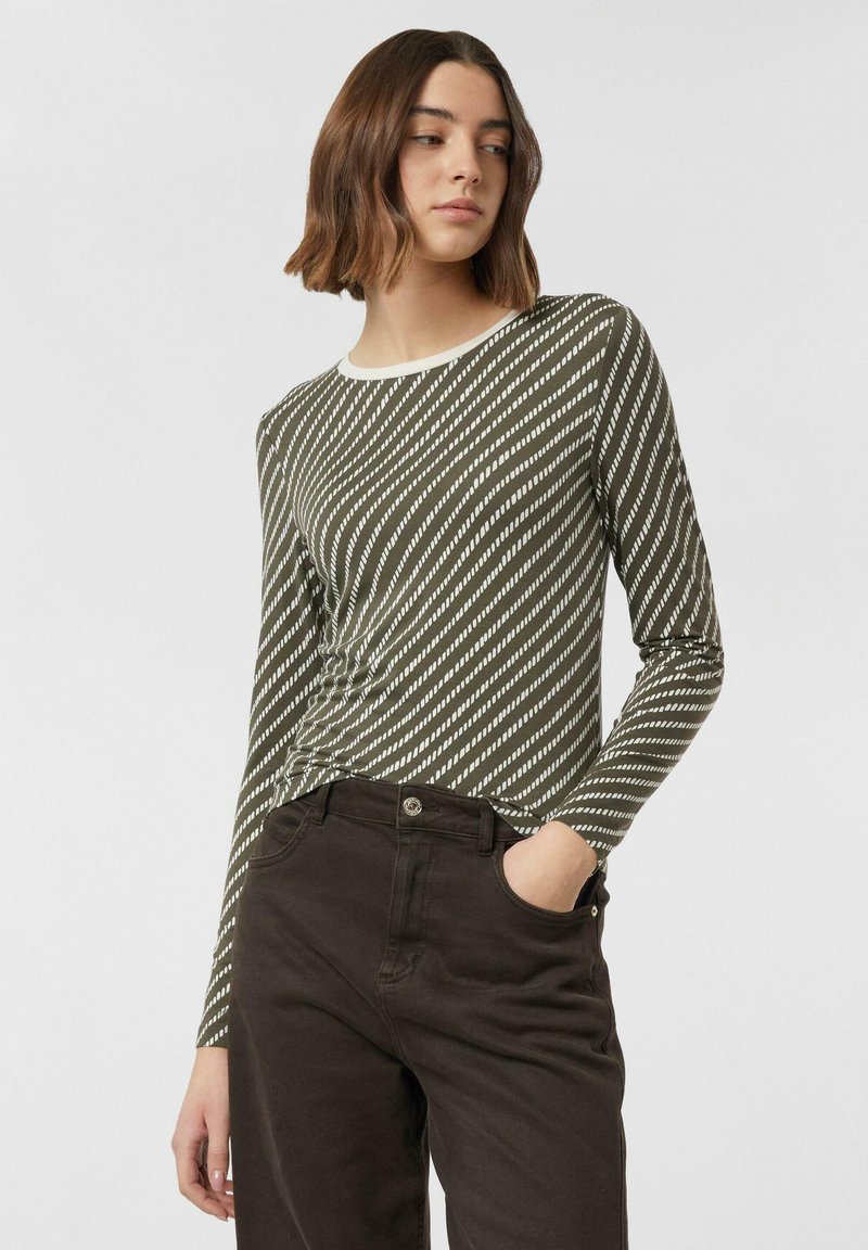 Junge Frau mit kurzen, welligen Haaren, die ein langärmeliges olivgrünes Shirt mit diagonalen weißen Streifen und schwarze Hosen trägt, eine Hand in der Tasche.