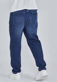 Tumesinised teksapüksid lõdva lõikega, millel on kaks tagataskut ja üleskeeratud ääred, kokku sobitatud valgete tossudega heledal taustal.