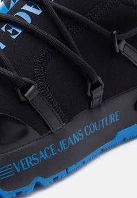 Μαύρο αθλητικό παπούτσι με μπλε λεπτομέρειες, υφή στο επάνω μέρος και σήμανση με το εμπορικό σήμα στη σόλα. Οι κορδόνια είναι ενσωματωμένα στο σχέδιο για ασφαλή εφαρμογή.