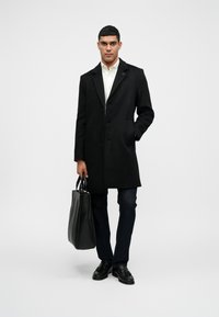 Sort uldfrakke med klapkrave, enkelt knaplukning, sidelommer. Sæt sammen med mørke jeans og sorte lædersko, mens han/hun holder en sort tote bag.