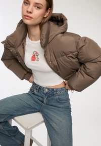 Brązowa kurtka puchowa z kapturem, z przednim zamkiem błyskawicznym i zatrzaskami. Biała dopasowana koszula z grafiką psa, w połączeniu z dopasowanymi niebieskimi dżinsami o wysokim stanie.