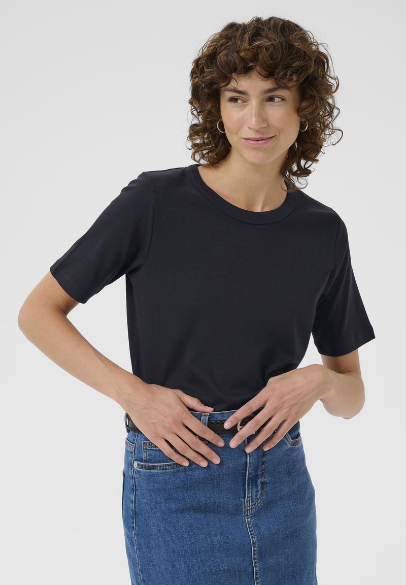 T-shirt noir à manches courtes avec col rond, en tissu doux, associé à une jupe en denim bleu. Le modèle se tient les mains sur les hanches.