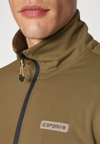 Olive green jacket with a high collar, black zipper, and Icepeak logo patch. Soft, textured fabric with a slim fit design.