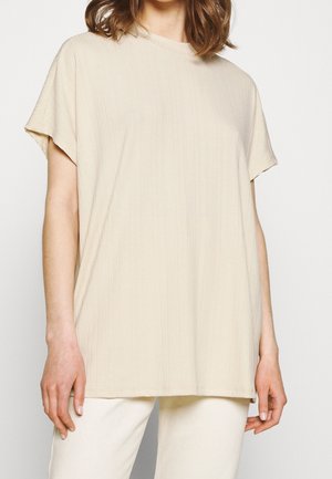 T-shirt beige a maniche corte in tessuto a coste con colletto a girocollo, vestibilità rilassata e orlo dritto, abbinato a pantaloni di colore chiaro.