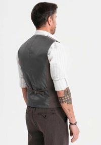 Muž s vyhrnutými rukávy a tetovanou pravou paží, oblečený ve fitted šedém vestě a odpovídajících kalhotách, otočený zády na bílém pozadí.