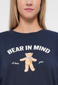 Navy blue sweatshirt featuring a textured graphic of a teddy bear with a blue scarf, and the text "BEAR IN MIND I love you" in white.