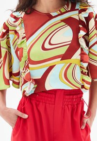 Blusa colorata con motivi vorticosi in rosso, verde, blu e crema, dotata di maniche a volant. Abbinata a pantaloni elastici in vita da un vivace rosso.