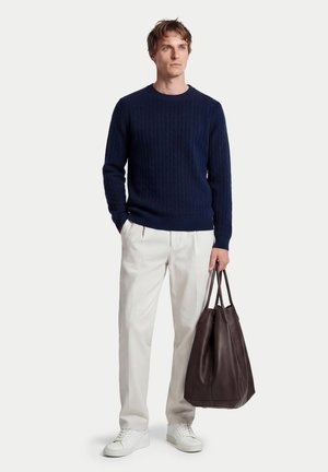 Mann trägt marineblauen Pullover, weiße Hose, weiße Turnschuhe, hält eine große dunkelbraune Ledertasche, steht vor einfarbigem Hintergrund.