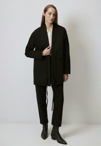 Giacca nera fino alla vita con maniche lunghe e due tasche frontali. Si annoda in vita; abbinata a pantaloni neri e stivaletti; texture del tessuto liscia.