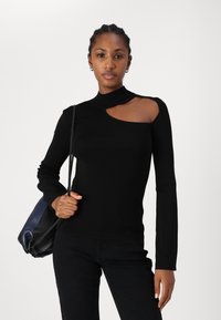 Haut noir à manches longues avec un col montant et une ouverture sur l'épaule. Le tissu est lisse et ajusté, associé à un pantalon sombre.
