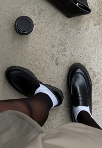 Mocassini in pelle nera con calzini bianchi e collant scuri su pavimento in cemento, accanto a una borsa nera e a una tazza di caffè con coperchio.