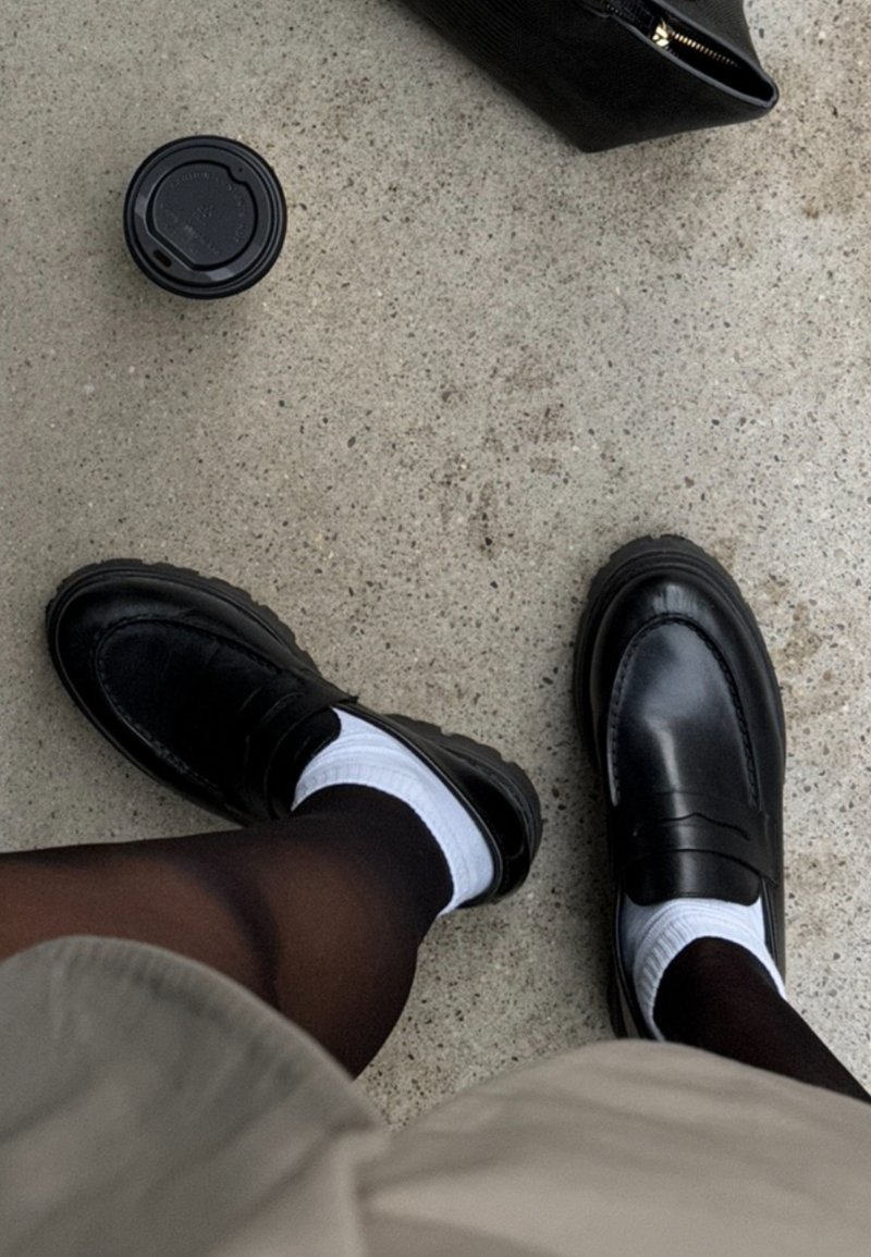 Mocassini in pelle nera con calzini bianchi e collant scuri su pavimento in cemento, accanto a una borsa nera e a una tazza di caffè con coperchio.