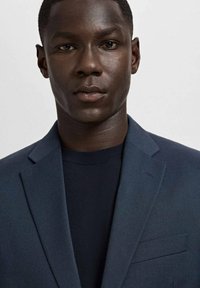 Młody czarnoskóry mężczyzna ubrany w ciemnoniebieską marynarkę i czarną bluzkę z okrągłym dekoltem, patrzący na wprost na jednolity jasny tle.