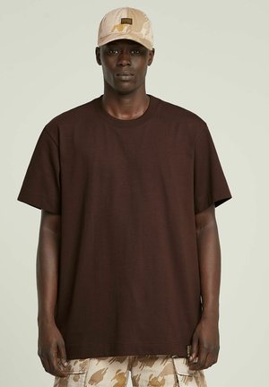 T-shirt marron à manches courtes avec une coupe décontractée, fabriqué en coton. Porté avec un short cargo à motif camouflage beige et une casquette assortie.