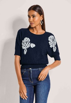 Vrouw draagt een marineblauwe, gestructureerde korte mouwen trui met witte rozenpatronen en donkerblauwe spijkerbroek, poserend tegen een eenvoudige lichte muur.