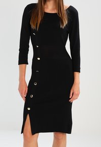Robe en maille noire avec manches trois-quarts, coupe ajustée, boutons sur le côté et fente haute sur la cuisse gauche.
