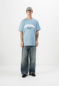 Ung mand iført en lyseblå Jack & Jones T-shirt, vide, lysnede jeans, sorte sneakers og en grå kasket, stående foran en hvid baggrund.