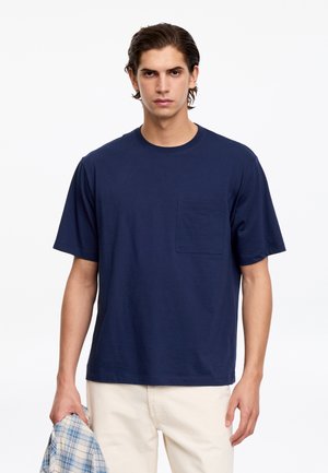 Jeune homme portant un T-shirt bleu marine avec une poche poitrine et un pantalon beige clair, tenant une chemise à carreaux bleue et blanche dans la main gauche.