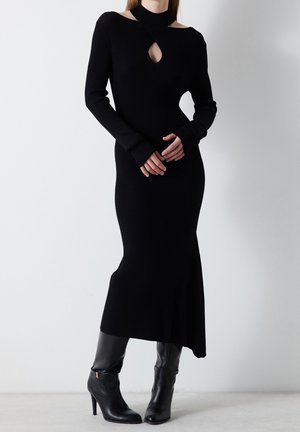 Femme portant une robe midi noire à manches longues avec des découpes au niveau du décolleté, associée à des bottes noires à talons hauts.