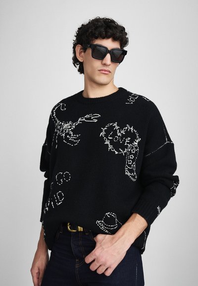 Pull noir en maille avec des motifs d'animaux et de chapeaux blancs, col rond, coupe décontractée, et poignets et ourlet côtelés. Porté avec un jean foncé.