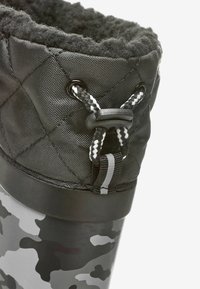 Botte étanche noire avec tige en tissu matelassé, doublure en fausse fourrure et semelle en caoutchouc à motif camouflage. Dispose de lacets noir et blanc avec un système de verrouillage.