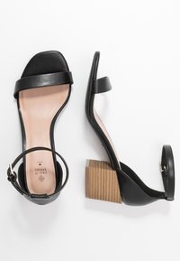 Sandales à talons en cuir noir avec un bout carré, une seule bride sur le pied, une bride à la cheville, et un talon bloc effilé avec une texture semblable au bois.