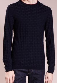 Homme portant un pull long à manches noires texturé avec un motif tressé et un jean bleu foncé sur un fond uni.