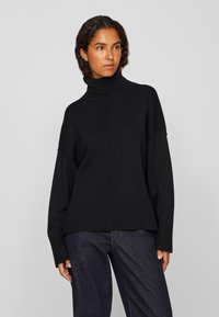 Μαύρο πουλόβερ με μανίκια σε άνετη εφαρμογή, ριγωτή υφή και φαρδιά μανίκια, συνδυασμένο με σκούρα τζιν παντελόνια.