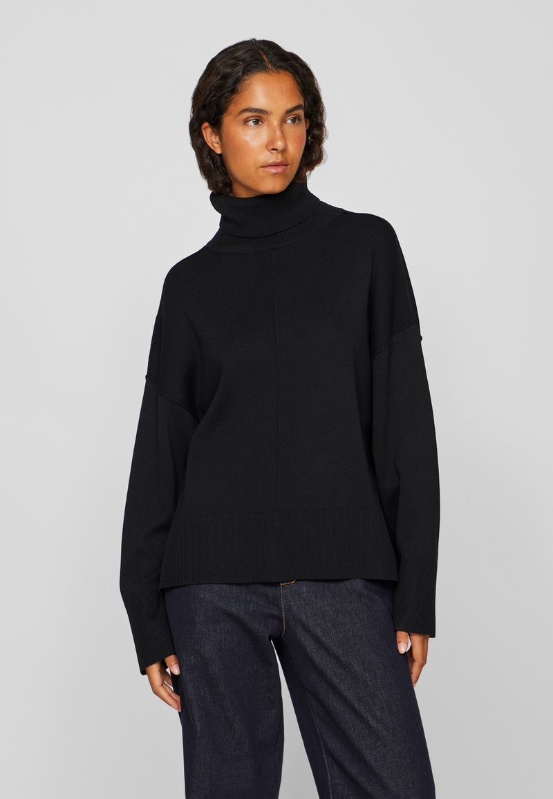 Μαύρο πουλόβερ με μανίκια σε άνετη εφαρμογή, ριγωτή υφή και φαρδιά μανίκια, συνδυασμένο με σκούρα τζιν παντελόνια.