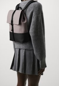 Mochila con parte superior de tela gris y base negra. Cuenta con un cierre de parte superior enrollable, correas ajustables y una superficie texturizada.