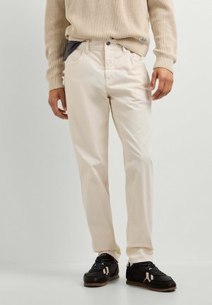Man wearing cream-colored pants, beige knit sweater, and black sneakers standing against a plain light background.