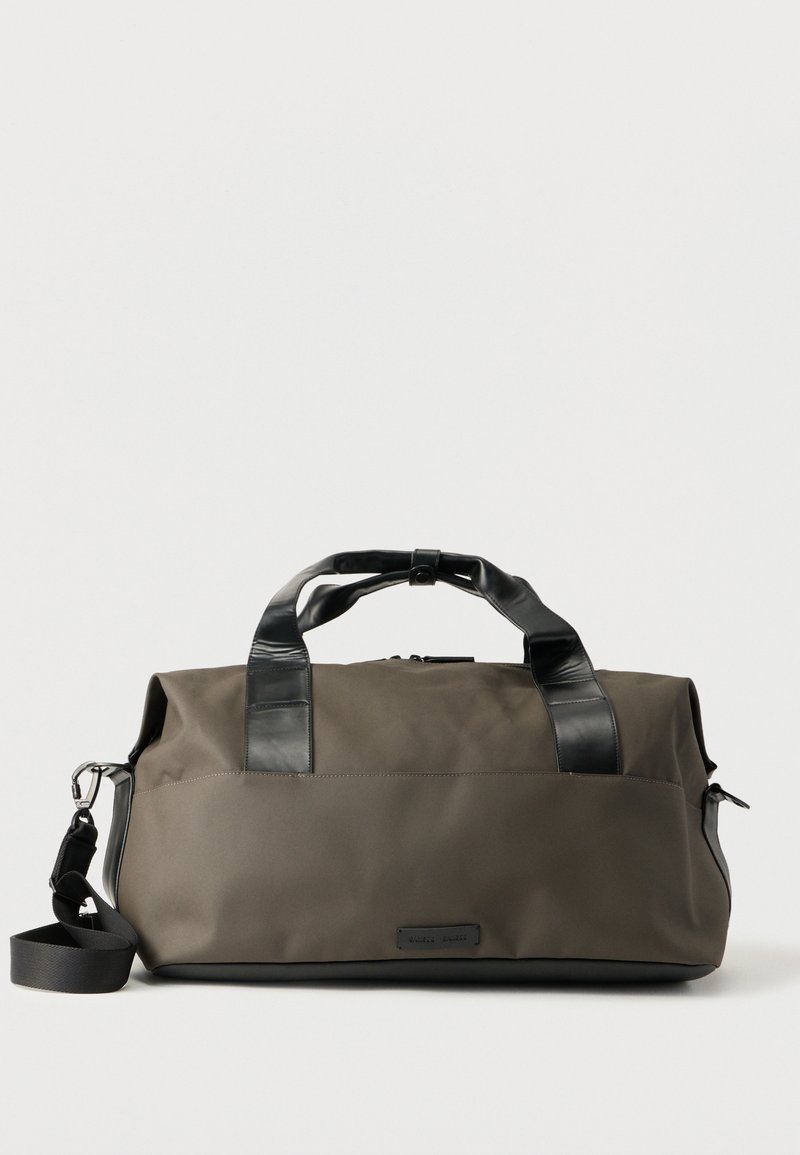 Τσαντάκι duffel σκούρου ελαιόχρωμου με μαύρες δερμάτινες λαβές και αποσπώμενο ιμάντα ώμου σε λευκό φόντο.
