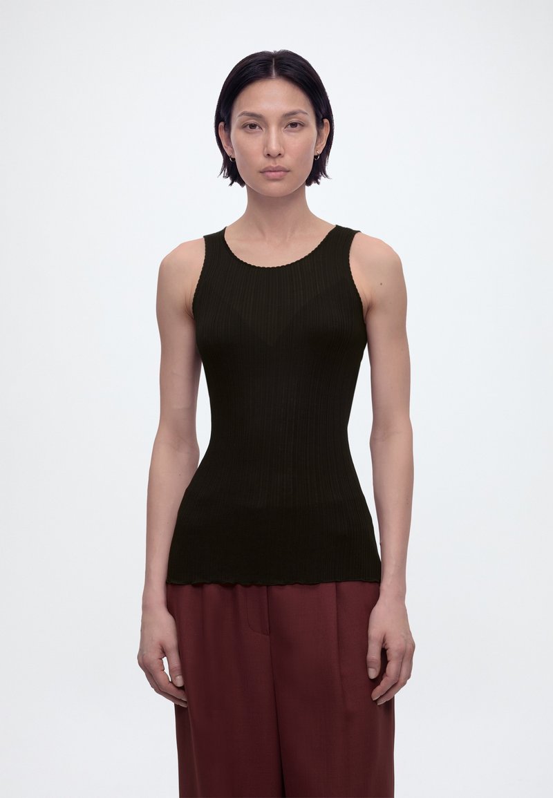 Γυναίκα με κοντά σκούρα μαλλιά που φοράει μαύρο αμάνικο ριμπ τοπ και φαρδιά μπορντό παντελόνια, στέκεται μπροστά σε απλό λευκό φόντο.