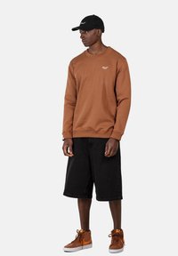 Sudadera marrón con puños acanalados, pantalones cortos de mezclilla negros y zapatillas altas marrones. Gorra negra con logotipo. El modelo está de pie sobre un fondo blanco.