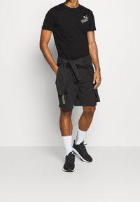 Camiseta negra de manga corta con un logo blanco de Puma, combinada con pantalones cortos negros. Unos calcetines blancos hasta el tobillo y zapatillas negras completan el conjunto.