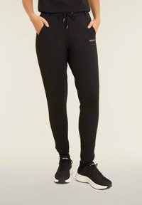 Pantalon de survêtement noir avec une coupe fuselée, taille à cordon, poches latérales et un petit logo doré sur la hanche gauche. Texture lisse.